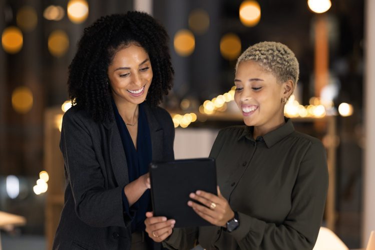 two women with dark colored clothes smiling in front of a tablet gadget